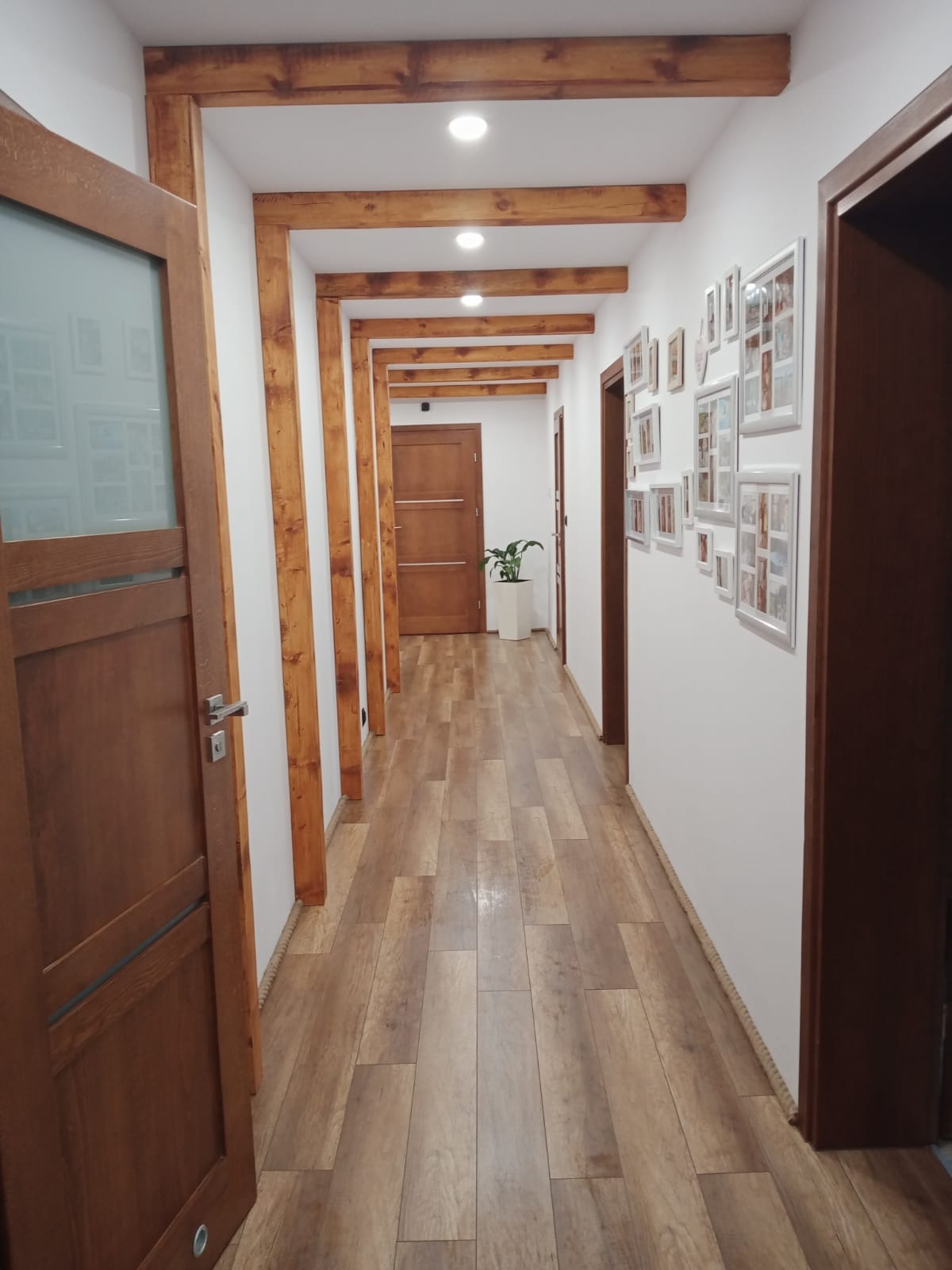 Długi, jasny korytarz z drewnianą podłogą, belkami stropowymi i ramkami na ścianie, wykończony w ciepłych brązach i bieli, z rośliną doniczkową w oddali.