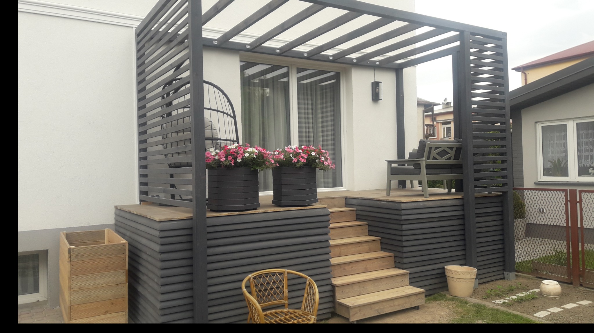 Zadaszony taras z drewnianą konstrukcją, schodami i meblami ogrodowymi, uzupełniony donicami z kwiatami.