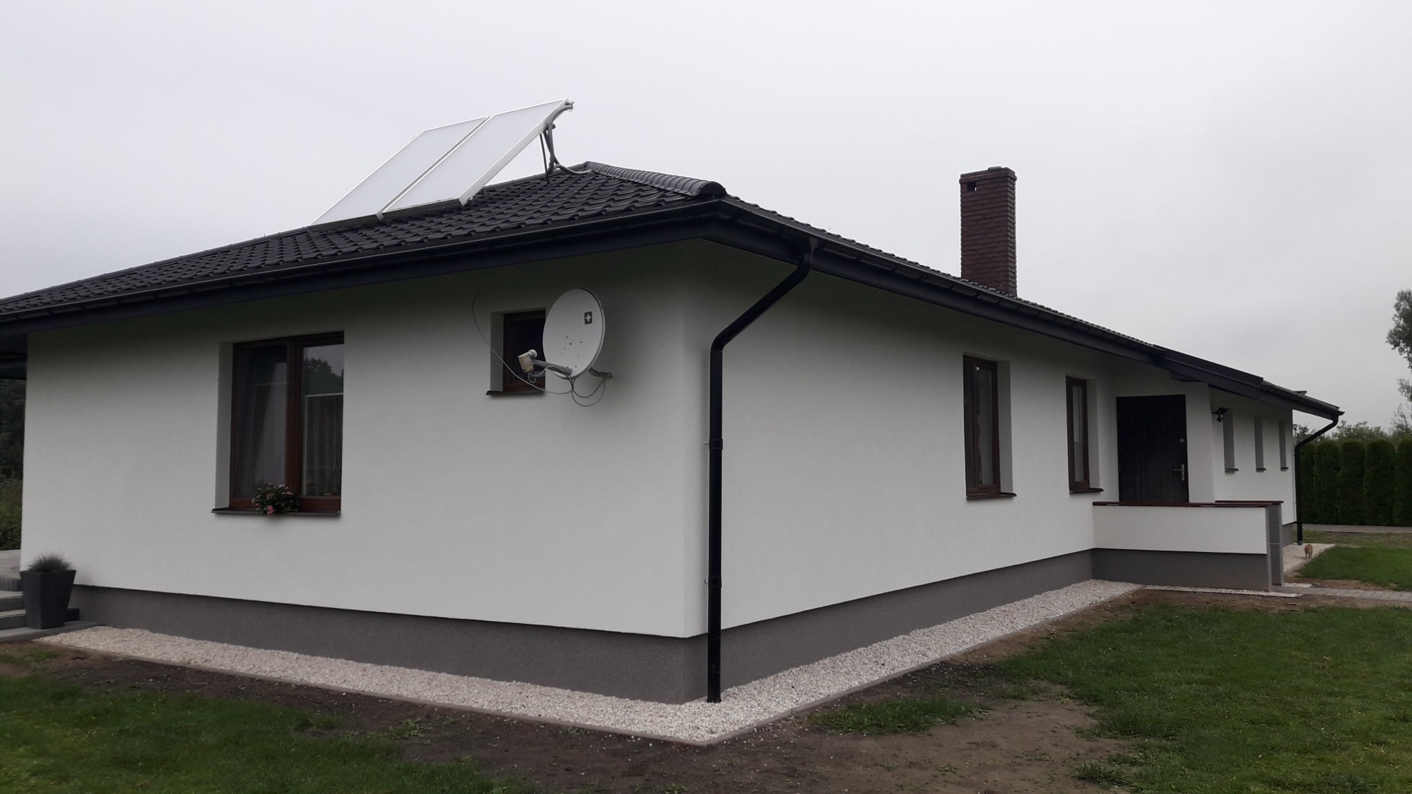 Biały, parterowy dom z ciemną dachówką i panelami słonecznymi, widoczna antena satelitarna na ścianie, szara podmurówka i białe kamyczki wzdłuż fundamentów.