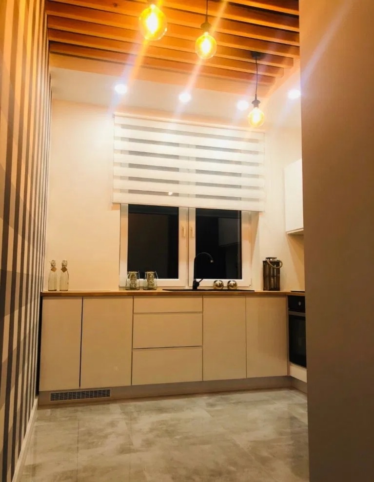 Nowoczesna kuchnia z beżowymi szafkami, drewnianym blatem, żaluzjami okiennymi w paski, dekoracyjnymi lampami wiszącymi i ścianą w czarno-białe pasy.