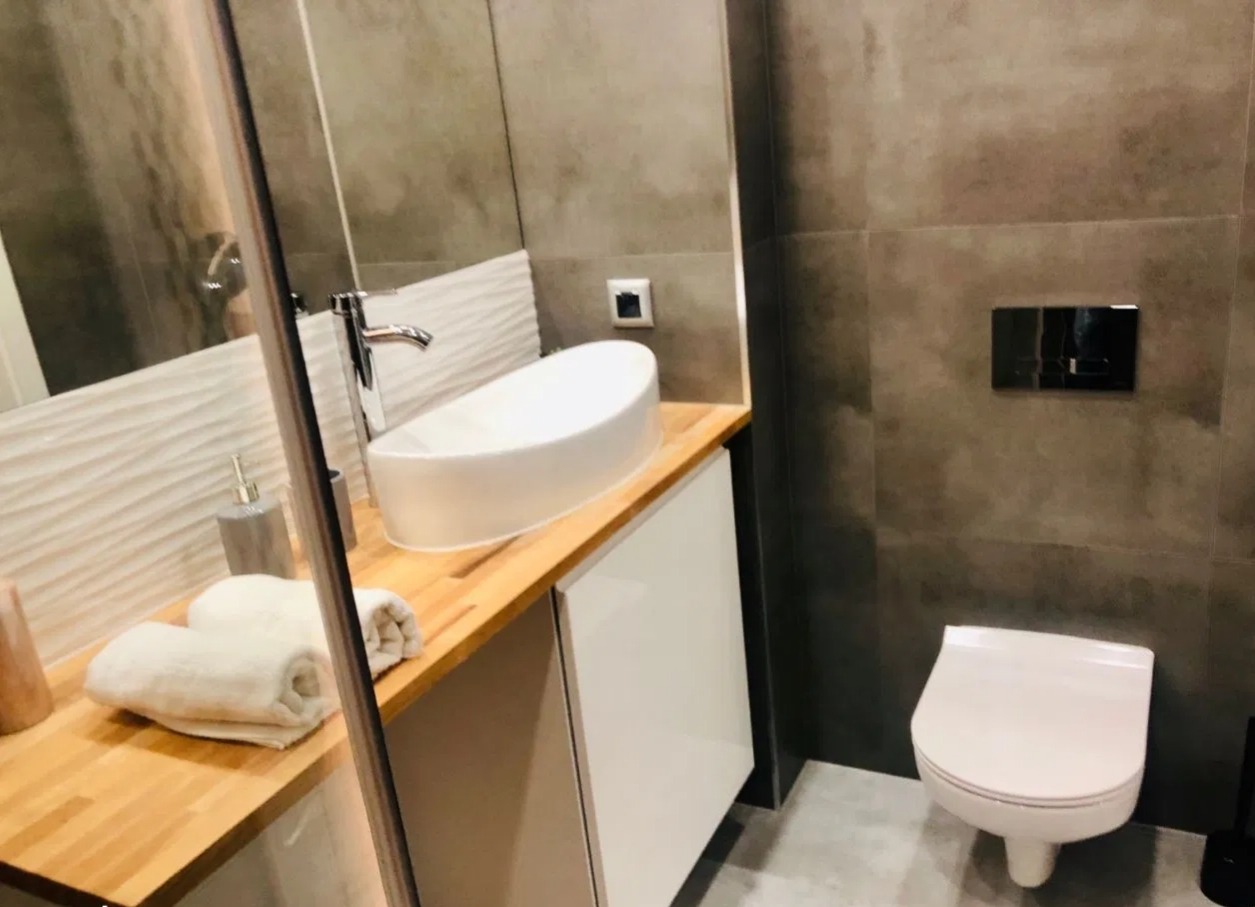 Nowoczesna łazienka z owalną umywalką nablatową, drewnianym blatem, białymi szafkami, szarą płytką ścienną i wiszącą toaletą.