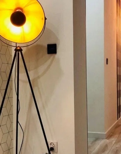 Czarny trójnożny reflektor z dużym, okrągłym kloszem oświetla fragment przedpokoju z geometryczną tapetą i pionowymi drewnianymi panelami na ścianie.