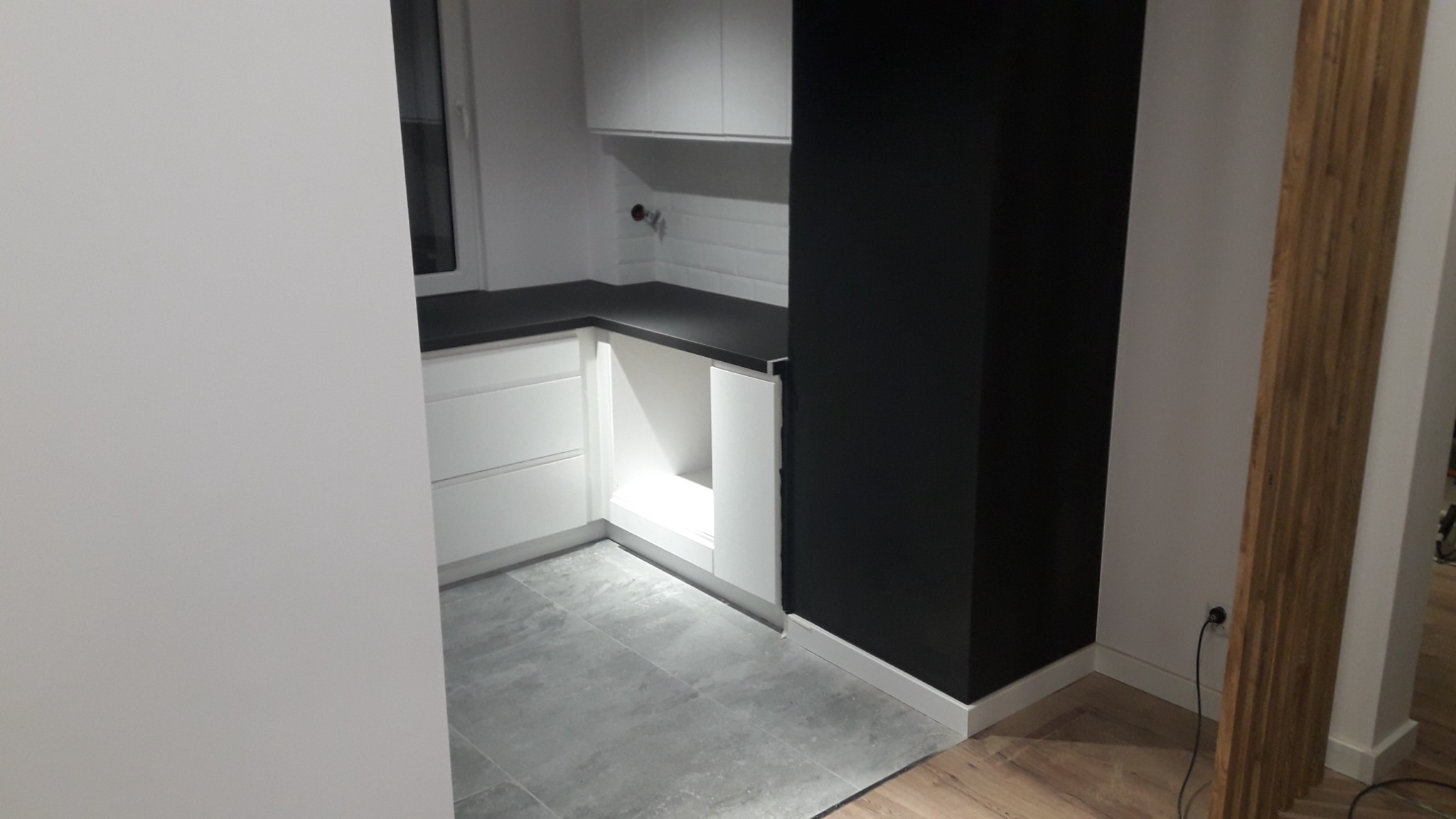 Nowoczesna kuchnia z białymi szafkami, czarnym blatem i ścianą, szarą podłogą oraz drewnianą dekoracją ściany w tle, widok z boku.