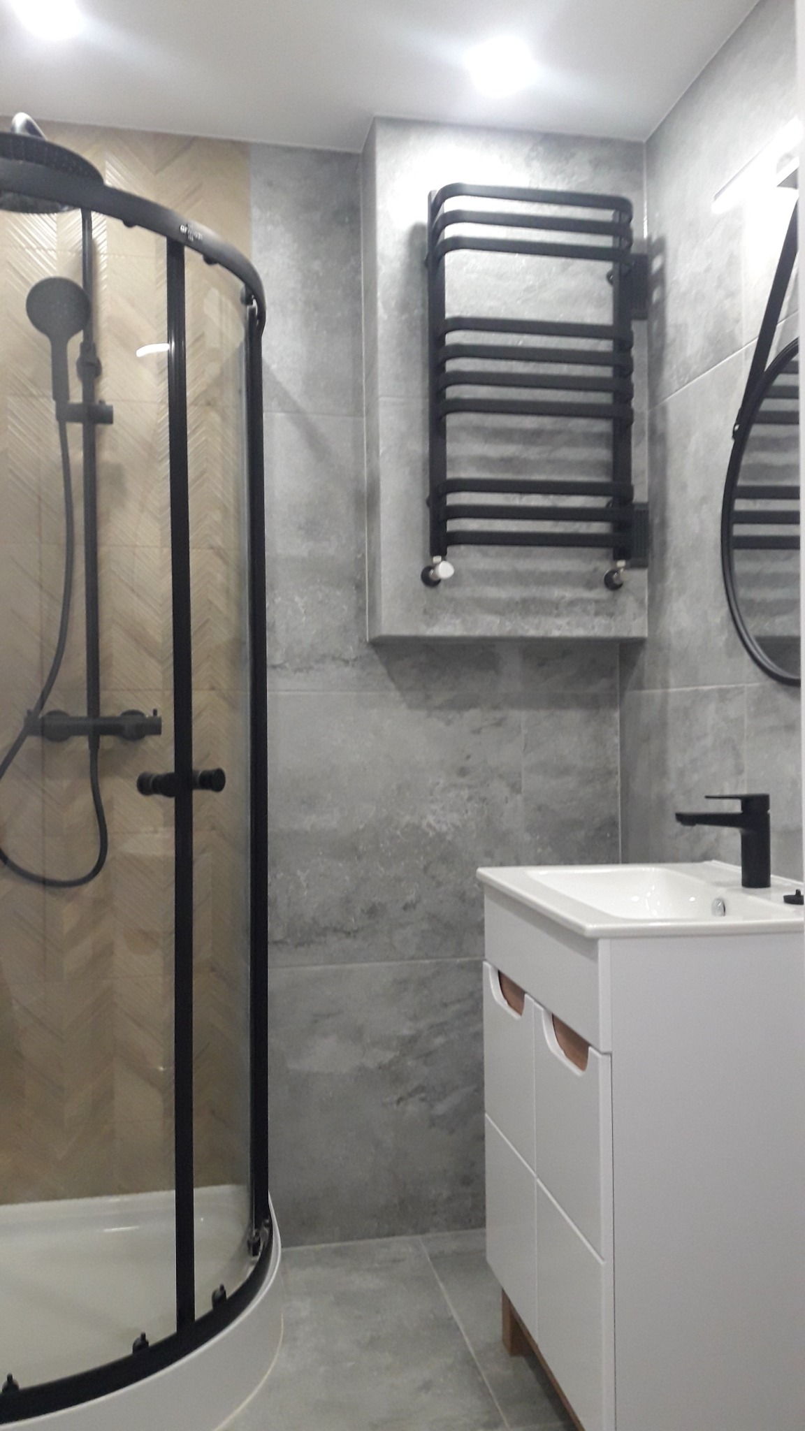 Nowoczesna łazienka z kabiną prysznicową z czarnymi profilami, grzejnikiem drabinkowym w kolorze czarnym i białą szafką umywalkową z szarymi płytkami na ścianach i podłodze.
