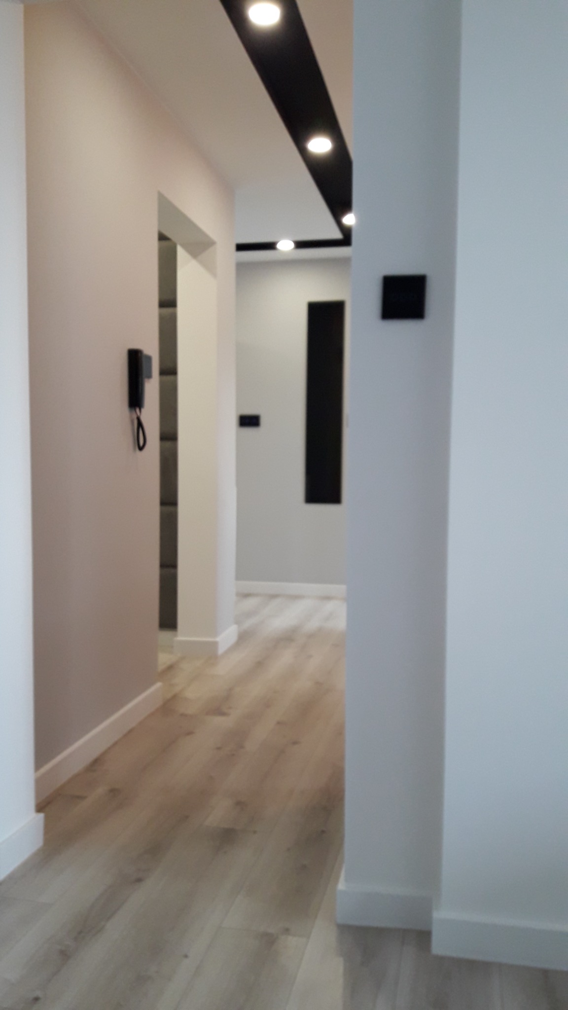 Nowoczesny korytarz z drewnopodobną podłogą, oświetleniem punktowym w czarnej listwie sufitowej i minimalistycznym interkomem na ścianie.