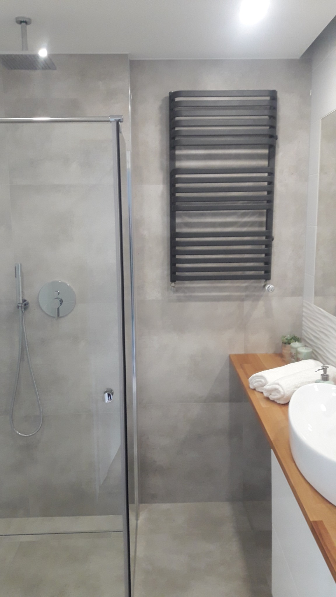 Nowoczesna łazienka z kabiną prysznicową typu walk-in, grzejnikiem drabinkowym na szarych płytkach i umywalką nablatową na drewnianym blacie.