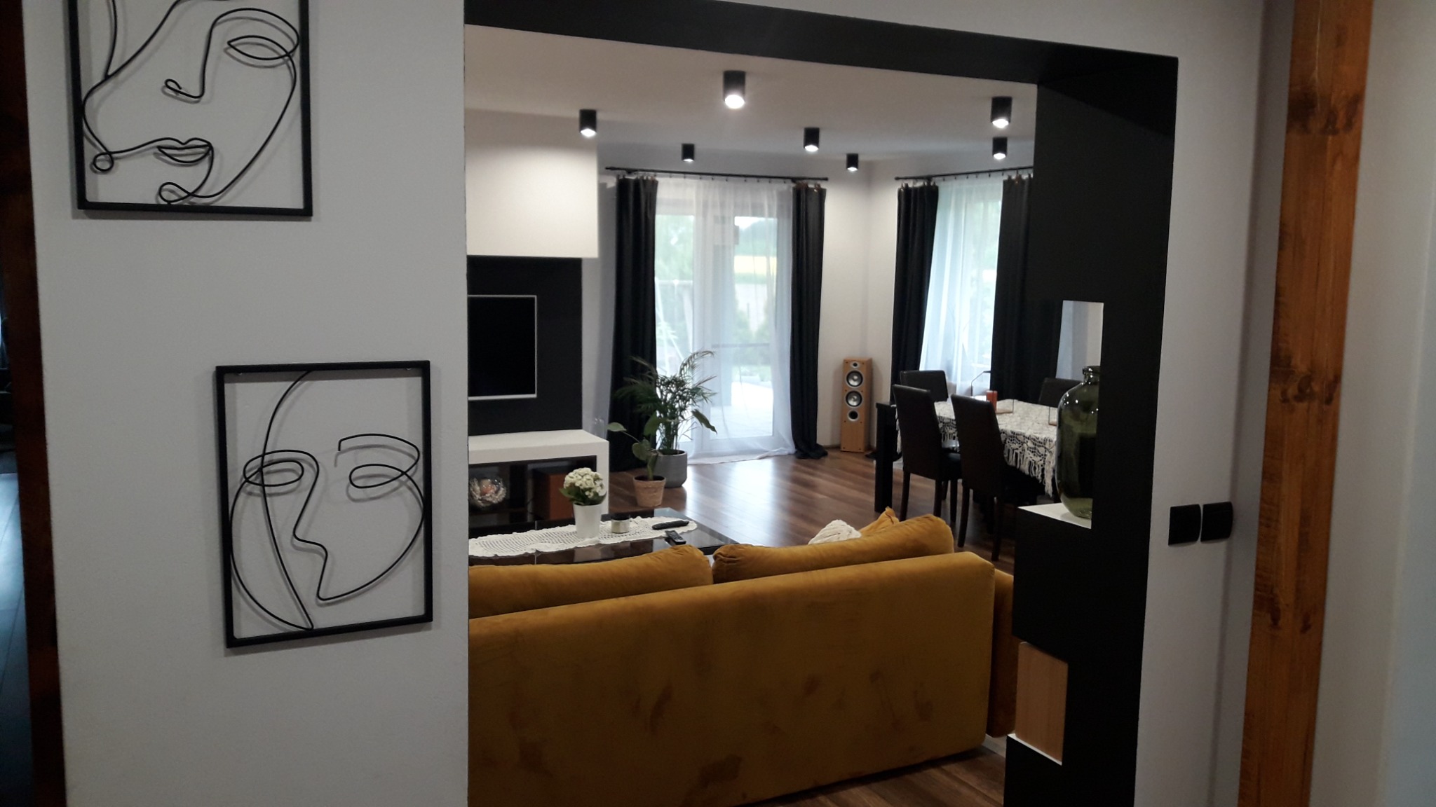 Nowoczesne wnętrze: salon z sofą w odcieniach musztardy, dekoracyjne grafiki na ścianie i widok na jadalnię z czarnymi zasłonami.