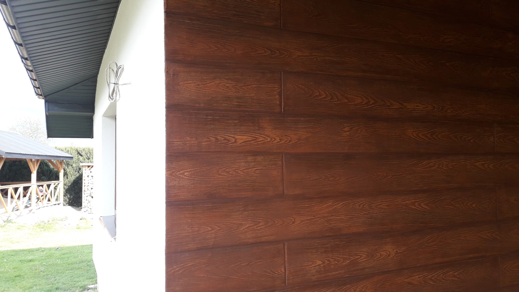 Elewacja budynku obłożona brązowymi panelami imitującymi drewno, widoczne fragmenty okna i dachu, w tle altana ogrodowa i ułożone drewno.