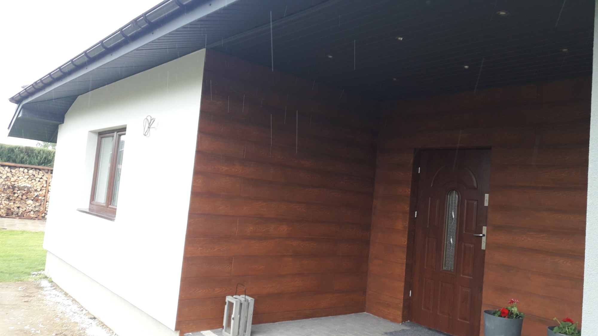 Elewacja budynku z drewnopodobną okładziną, brązowe drzwi wejściowe, biała ściana z oknem, dekoracyjna latarenka i donice z czerwonymi kwiatami przed wejściem.