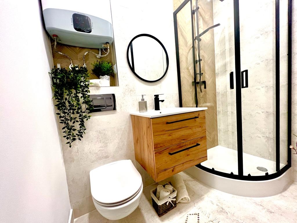 Nowoczesna łazienka z wiszącą szafką z drewnianymi frontami, okrągłym lustrem, kabiną prysznicową z czarnymi ramami i wiszącą toaletą. Nad toaletą zamontowany bojler i dekoracyjne rośliny.