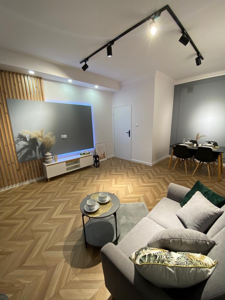 Nowoczesne, jasne wnętrze biura z parkietem w jodełkę, szarą sofą, stolikiem kawowym z filiżankami, ścianą TV z drewnianymi lamelami i oświetleniem punktowym.