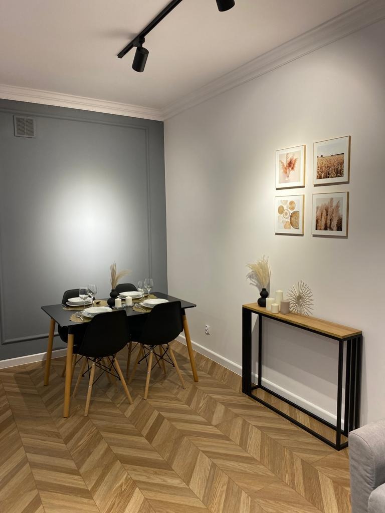 Nowoczesne wnętrze biurowe z drewnianą podłogą w jodełkę, czarnym stołem i krzesłami, szarą ścianą z dekoracyjną sztukaterią oraz minimalistyczną konsolą pod ścianą z galerią obrazków.