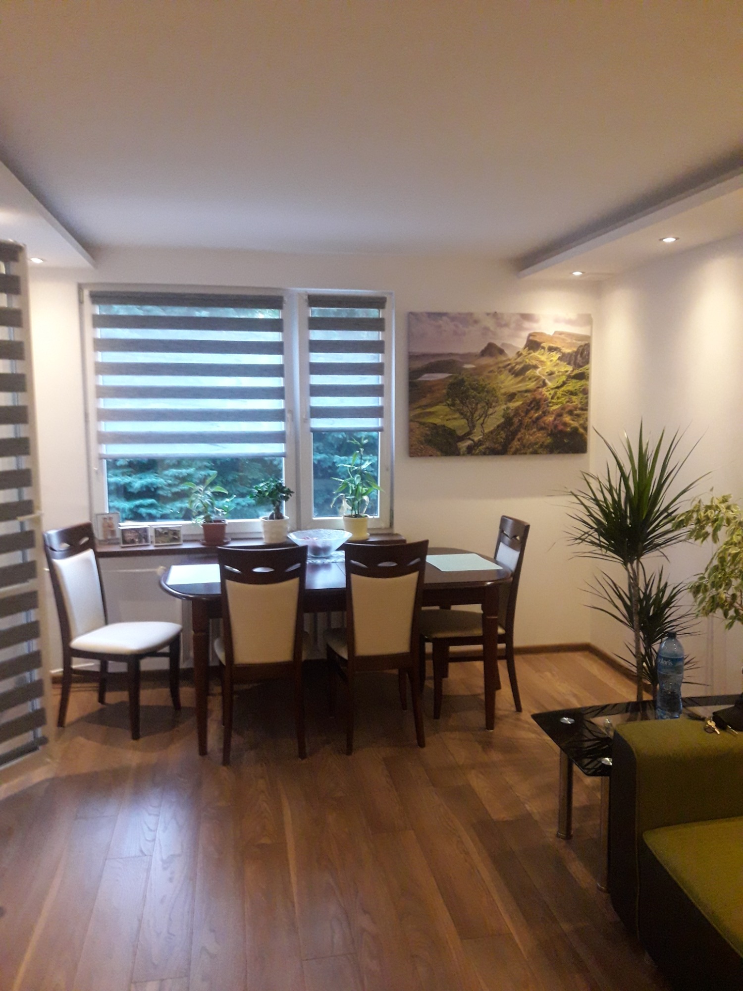 Jasna jadalnia z drewnianym stołem, krzesłami z kremowym obiciem, parkietem, roletami dzień-noc i obrazem górskim na ścianie.