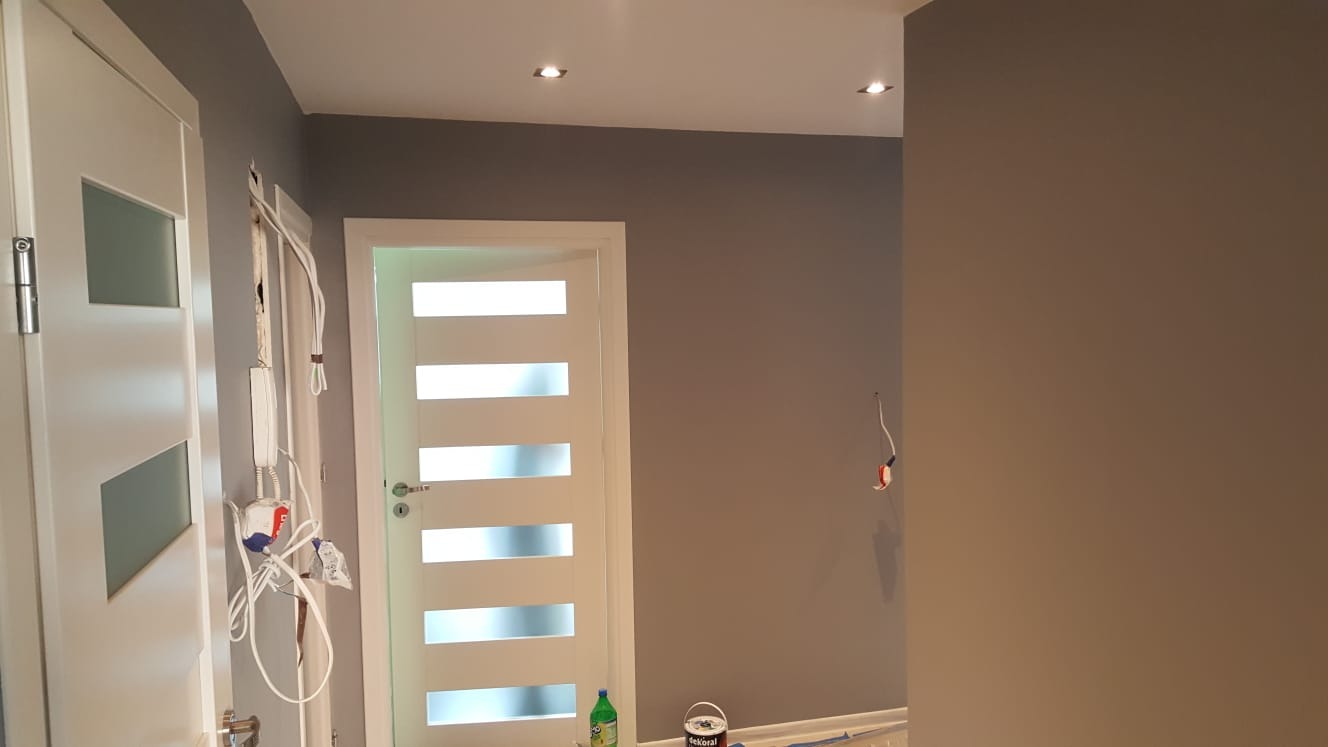 Świeżo pomalowane szare ściany w korytarzu, widoczne dwie białe drzwi, puszka farby Dekoral na podłodze i wystające kable elektryczne.