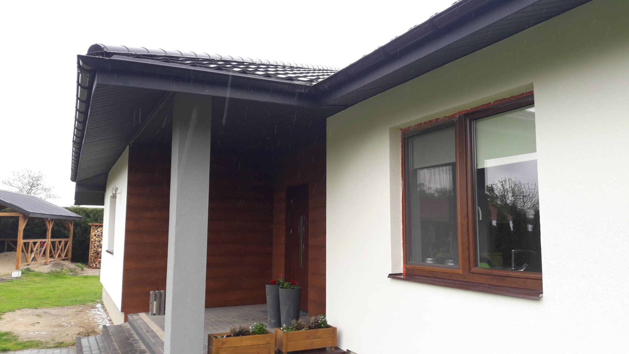 Elewacja domu jednorodzinnego z oknem w drewnianej ramie, wykończona białą gładzią i drewnianą okładziną, widok na zadaszone wejście z kolumnami i czarnym dachem, w tle drewniana altana.