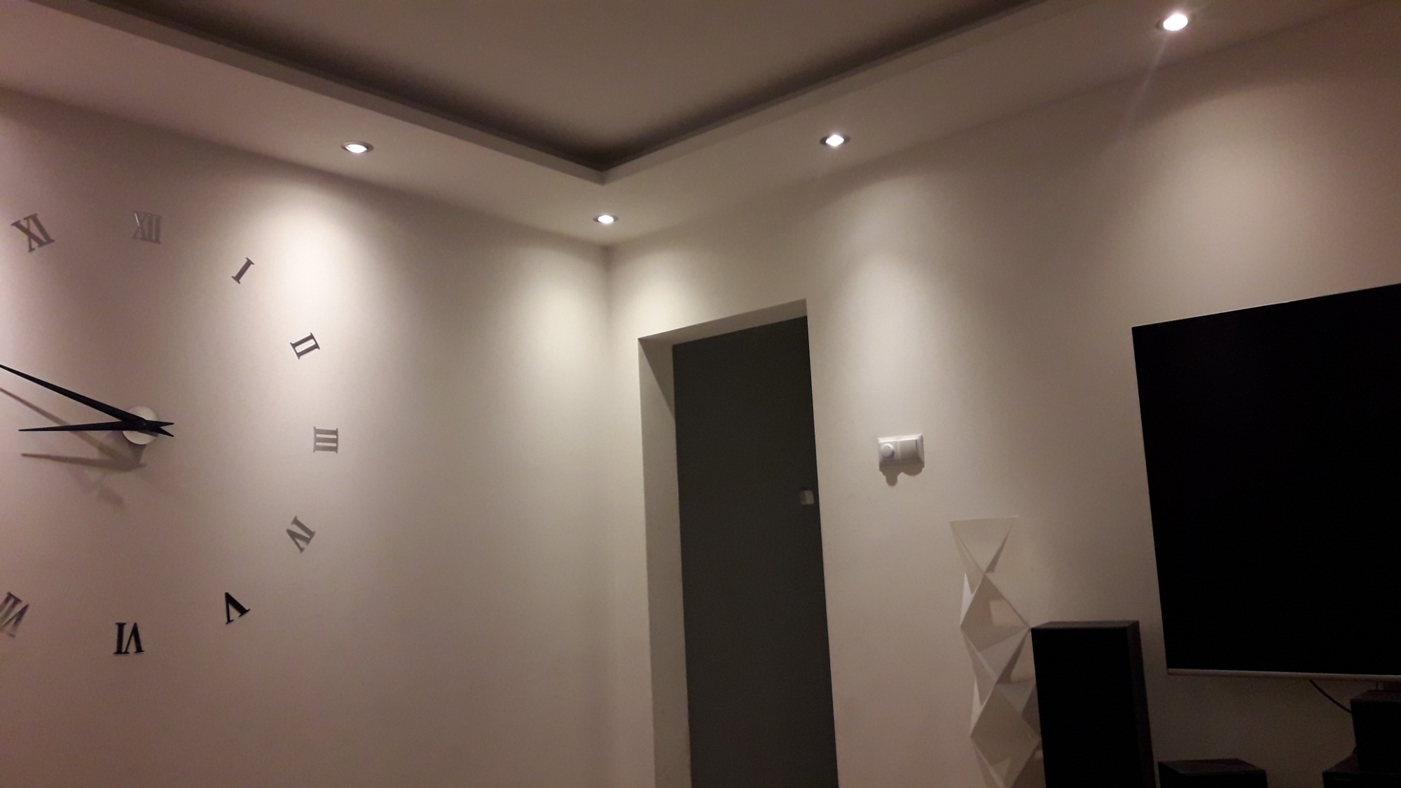 Nowoczesne wnętrze z białymi ścianami, podwieszanym sufitem z punktowym oświetleniem, dużym zegarem ściennym z rzymskimi cyframi, ciemnymi drzwiami i elementami dekoracyjnymi.