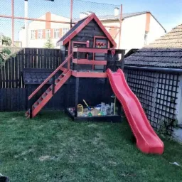 Drewniany domek dla dzieci z czerwoną zjeżdżalnią i piaskownicą na trawniku, widoczny z boku, z elementami czarnego i czerwonego malowania.