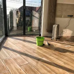 Ułożona podłoga z paneli imitujących drewno w jasnym pomieszczeniu z dużymi oknami, widoczne wiadro i narzędzia, fragment ściany z grzejnikiem i gniazdkami.