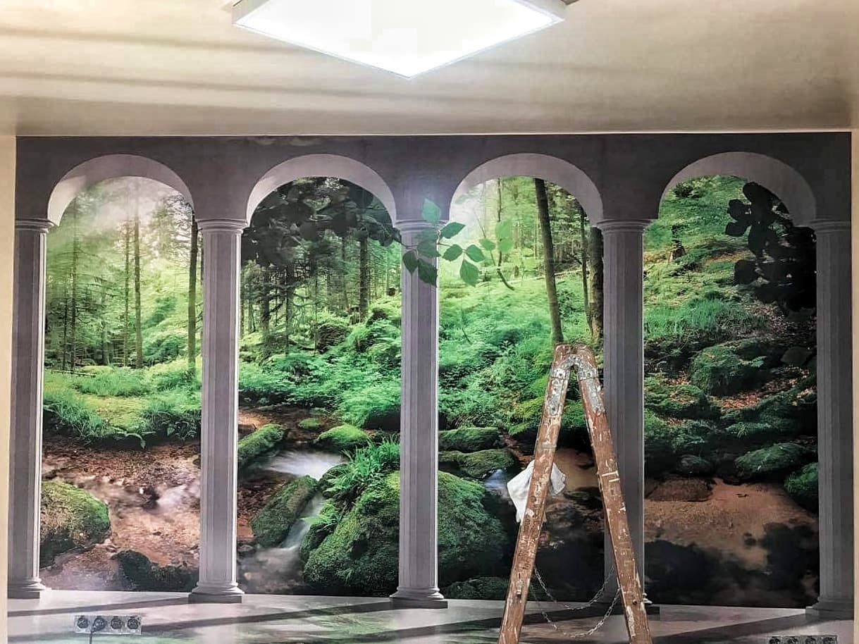 Malowanie ściany z fototapetą imitującą widok na leśny strumień przez kamienne kolumny; drabina oparta o ścianę, widoczne gniazdka elektryczne przy podłodze.
