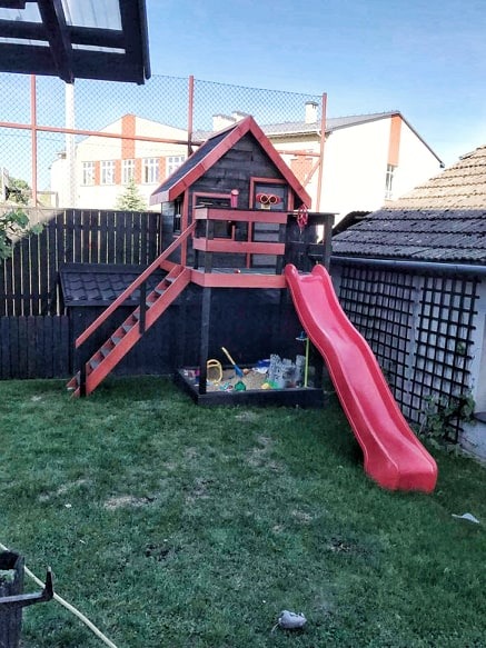 Drewniany domek dla dzieci z czerwoną zjeżdżalnią i piaskownicą na trawniku, widoczny z boku, z elementami czarnego i czerwonego malowania.