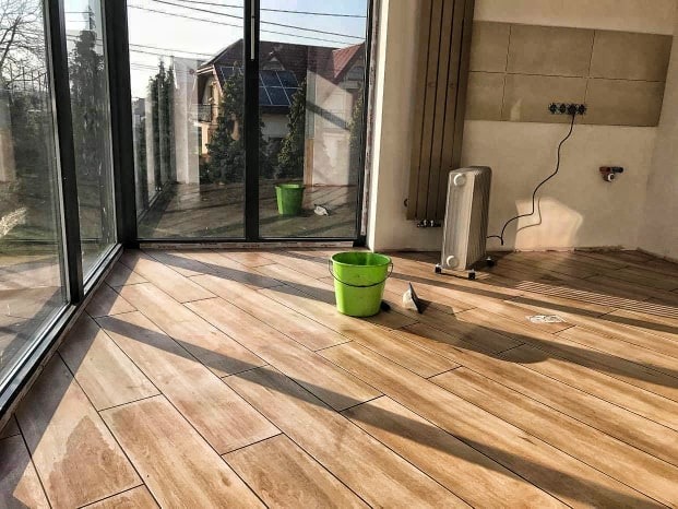 Ułożona podłoga z paneli imitujących drewno w jasnym pomieszczeniu z dużymi oknami, widoczne wiadro i narzędzia, fragment ściany z grzejnikiem i gniazdkami.