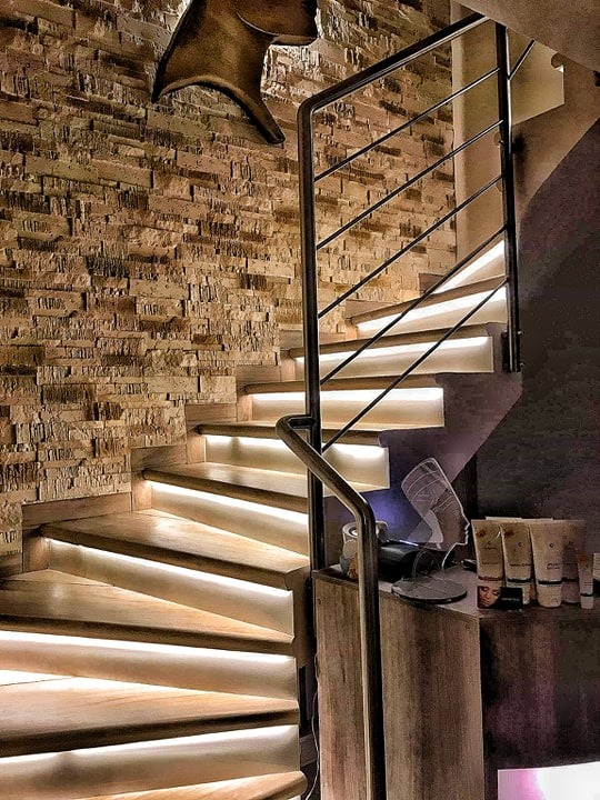 Spiralne schody z drewnianymi stopniami podświetlonymi paskami LED, ściana wyłożona kamieniem dekoracyjnym, metalowa balustrada, dekoracja w kształcie głowy zwierzęcia na ścianie, kosmetyki...