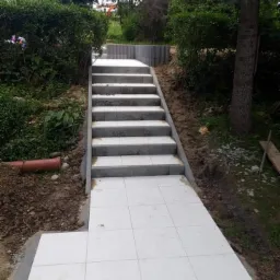 Ułożone białe płytki chodnikowe prowadzące do schodów z szarymi obrzeżami, otoczone zielenią i ziemią, z widocznym fragmentem pomarańczowej rury.