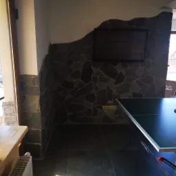 Wnętrze pokoju z kamienną ścianą o nieregularnym kształcie, na której wisi telewizor. Pod ścianą stoi stół do ping-ponga marki Kettler. Podłoga wyłożona ciemnymi płytkami. Widoczny fragment...