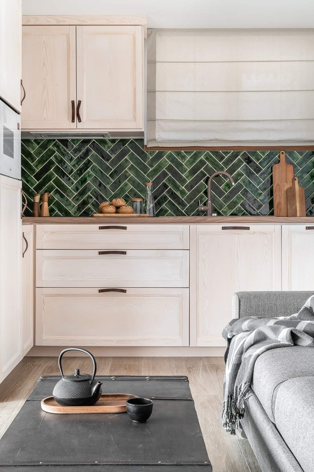 Jasna kuchnia z zielonymi płytkami ułożonymi w jodełkę na ścianie, drewnianym blatem i białymi szafkami z ciemnobrązowymi uchwytami. Na blacie leżą chleby, przyprawy i deski do krojenia.