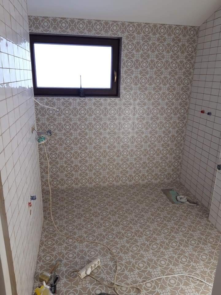 Łazienka w trakcie remontu: ściany częściowo wyłożone białymi kwadratowymi płytkami, pozostałe ściany i podłoga pokryte wzorzystą tapetą w odcieniach beżu i bieli, widoczne gniazdko elektryczne...