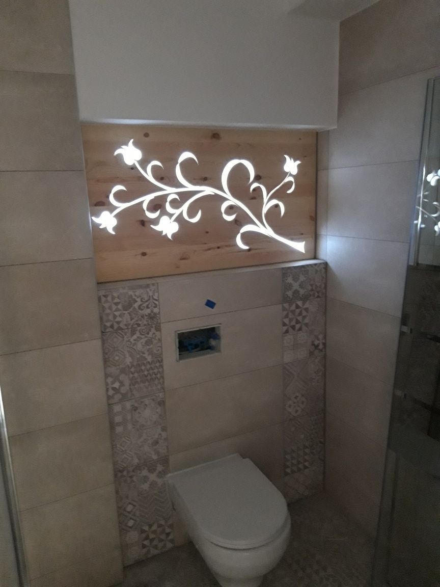 Wykończona toaleta z podwieszaną muszlą klozetową, obudowana beżowymi płytkami z dekoracyjnymi wstawkami i podświetlanym drewnianym panelem z motywem roślinnym.