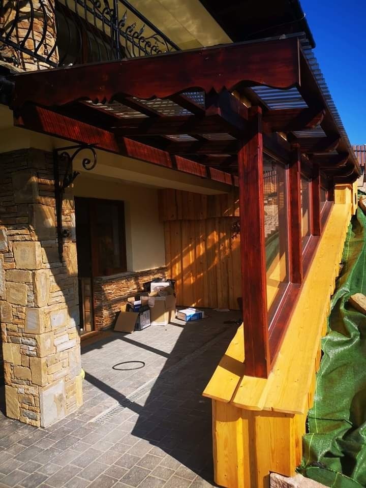 Zadaszenie tarasu z drewnianą konstrukcją w kolorze ciemnego brązu, wsparte na kamiennej kolumnie z dekoracyjnym metalowym wspornikiem, kostka brukowa na posadzce tarasu, widoczne kartony i resztki...