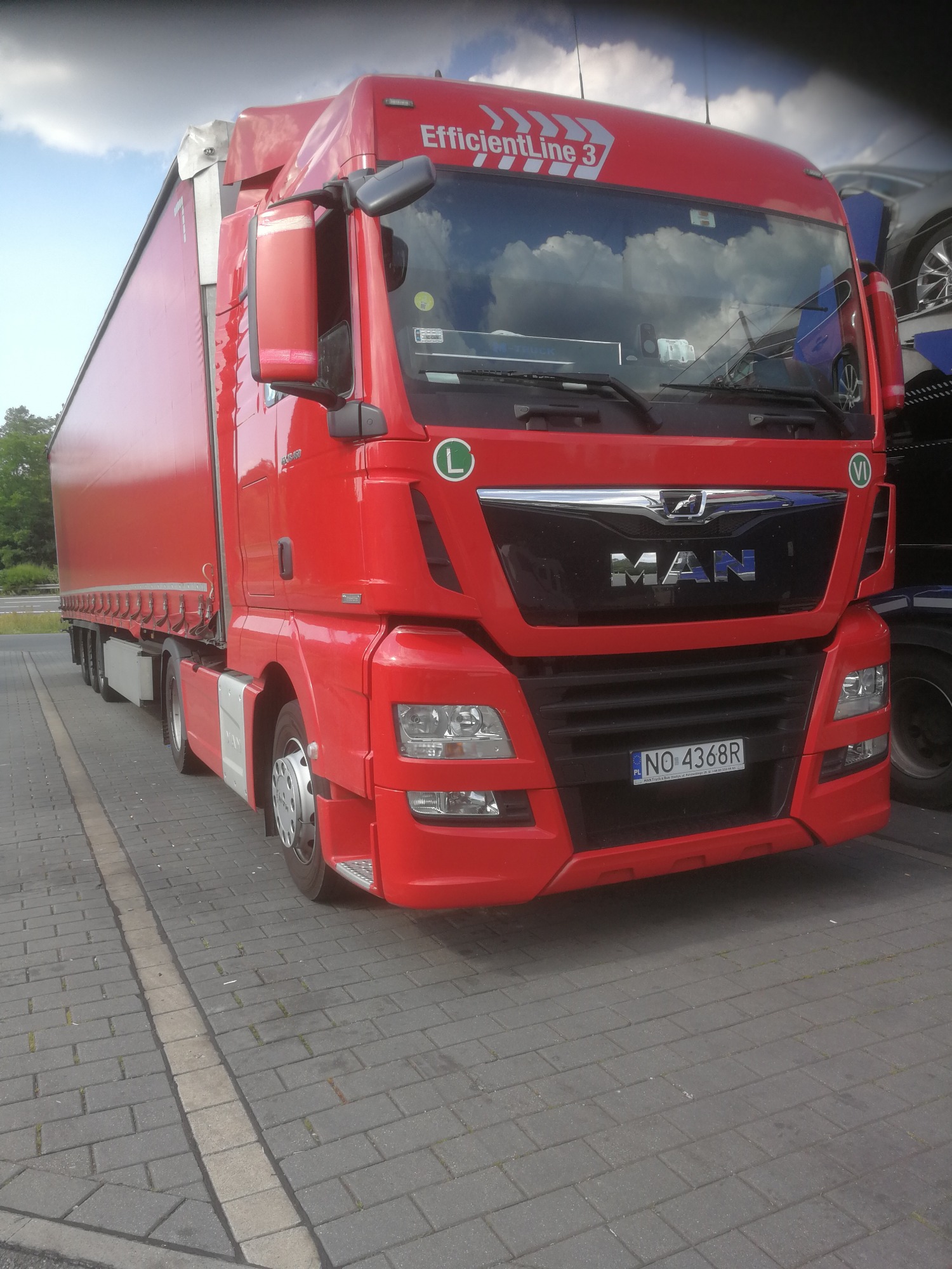 Czerwona ciężarówka MAN EfficientLine 3 z naczepą, zaparkowana na brukowanym parkingu, z widoczną tablicą rejestracyjną NO-4368R.