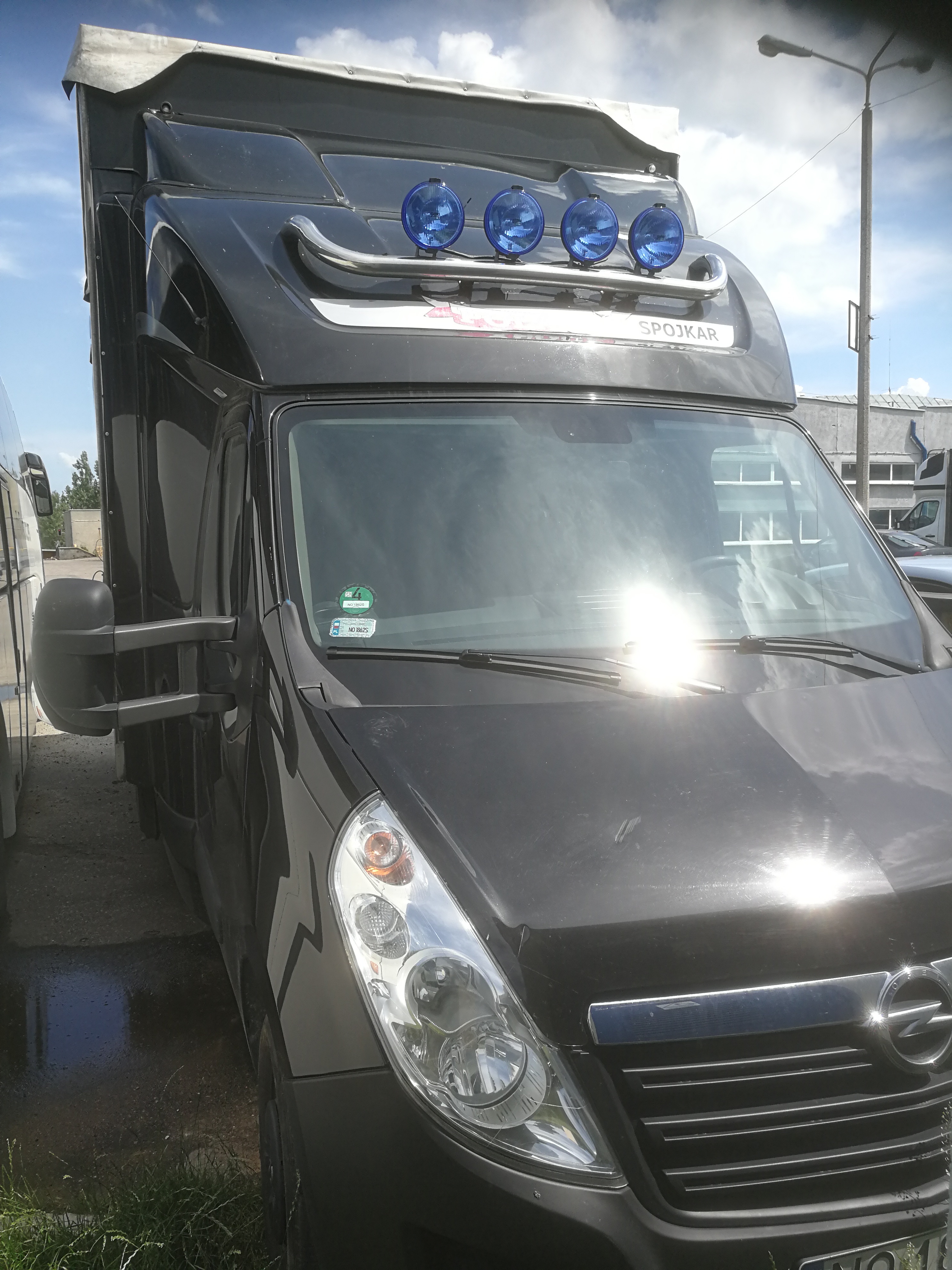 Czarny Opel Movano z wysokim dachem i dodatkowymi niebieskimi reflektorami na dachu, widok z przodu, z naklejką 'SPOJKAR'.