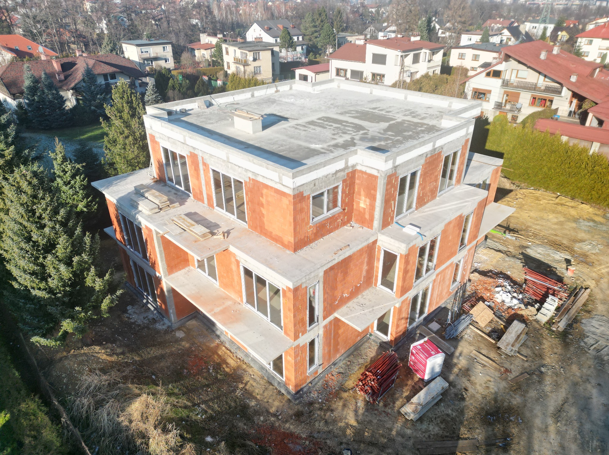 Dwukondygnacyjny dom w stanie surowym otwartym, z czerwonej cegły i białego betonu, widok z góry na tle zabudowy jednorodzinnej i drzew. Na ziemi leżą materiały budowlane.