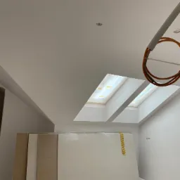 Poddasze w trakcie remontu: widoczne płyty gotowe do montażu, dwa okna dachowe wpuszczające jasne światło, świeżo pomalowane ściany i wystające przewody instalacji elektrycznej.