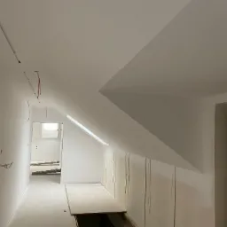 Poddasze w trakcie remontu: białe ściany, skośny sufit, widoczne przewody elektryczne, fragment otworu w podłodze, zabezpieczone folią drzwi.