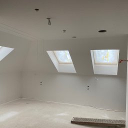 Łukasz Prace Wykończeniowe - Poddasze w trakcie prac wykończeniowych, widoczne trzy okna dachowe, gładkie, białe ściany i sufit z wystającymi przewodami elektrycznymi, na podłodze leży metalowa kratka.