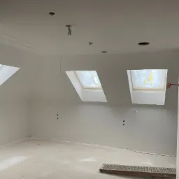 Poddasze w trakcie prac wykończeniowych, widoczne trzy okna dachowe, gładkie, białe ściany i sufit z wystającymi przewodami elektrycznymi, na podłodze leży metalowa kratka.