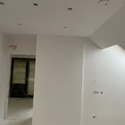 Wnętrze w trakcie malowania na biało, widoczne otwory na oświetlenie w suficie, niezamaskowane gniazdka elektryczne w ścianie, drzwi zabezpieczone folią ochronną.