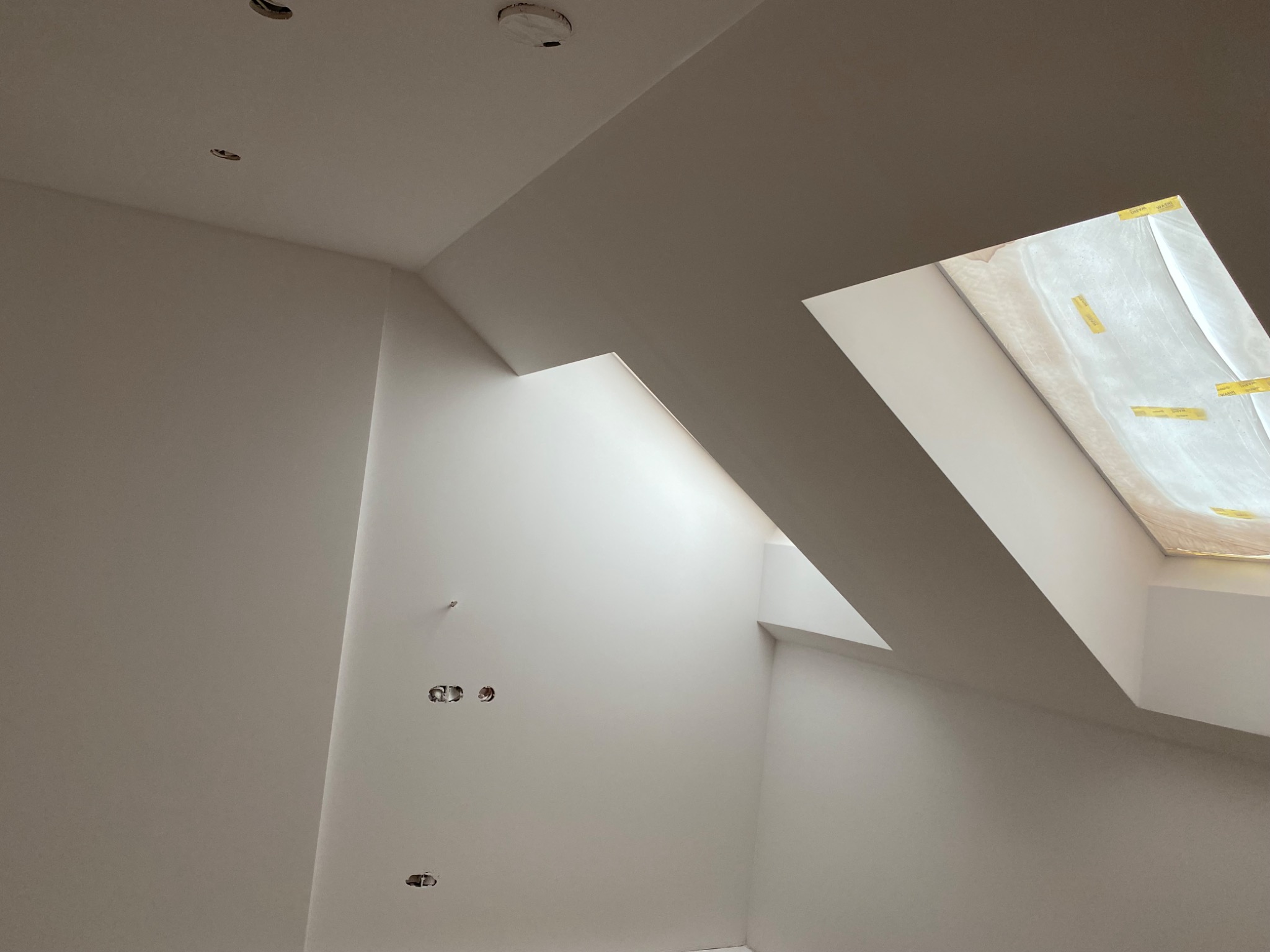 Wnętrze poddasza po świeżym pomalowaniu na biało, widoczne gniazdka elektryczne bez osprzętu i okno dachowe zasłonięte folią ochronną.