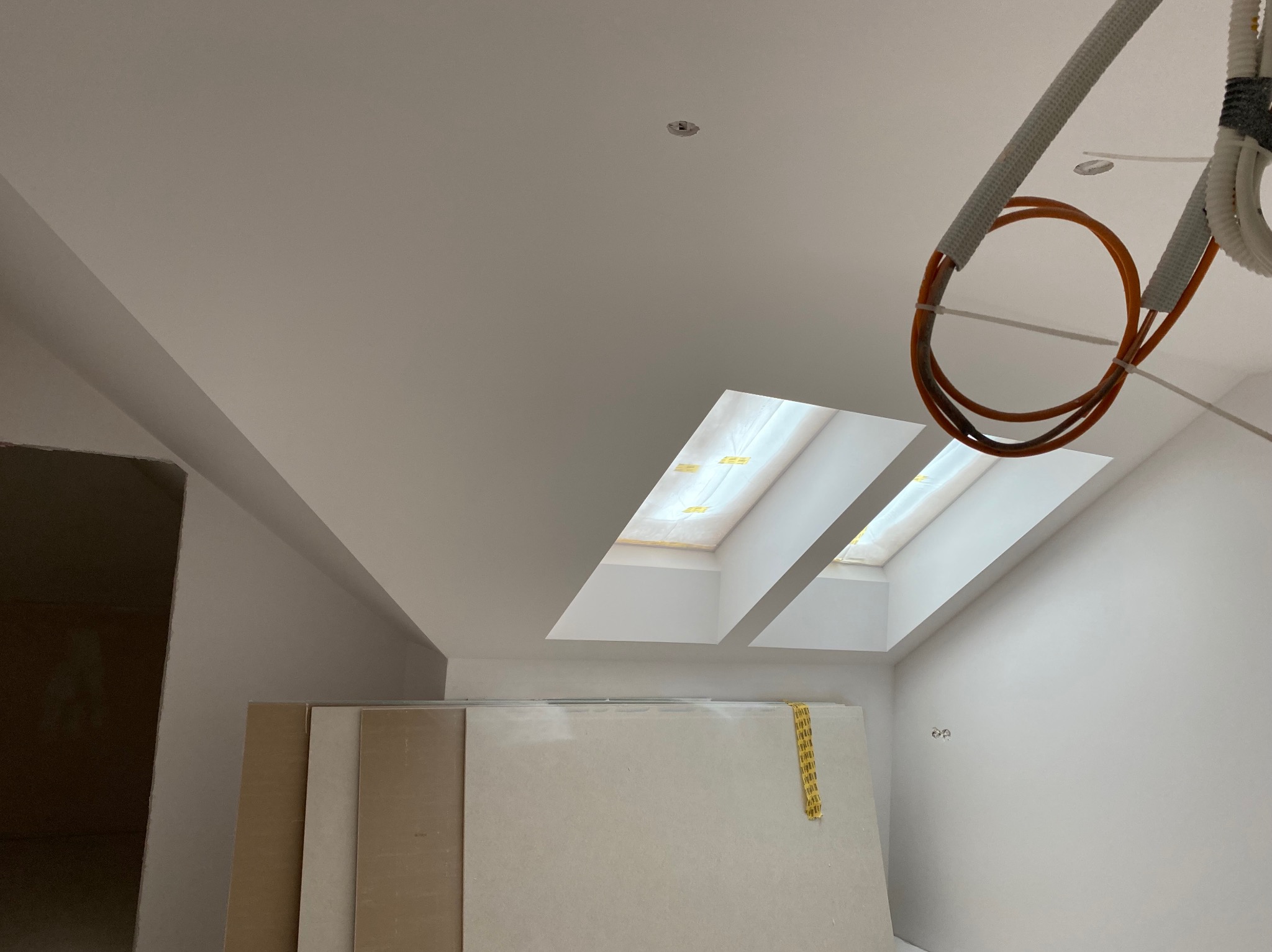 Poddasze w trakcie remontu: widoczne płyty gotowe do montażu, dwa okna dachowe wpuszczające jasne światło, świeżo pomalowane ściany i wystające przewody instalacji elektrycznej.