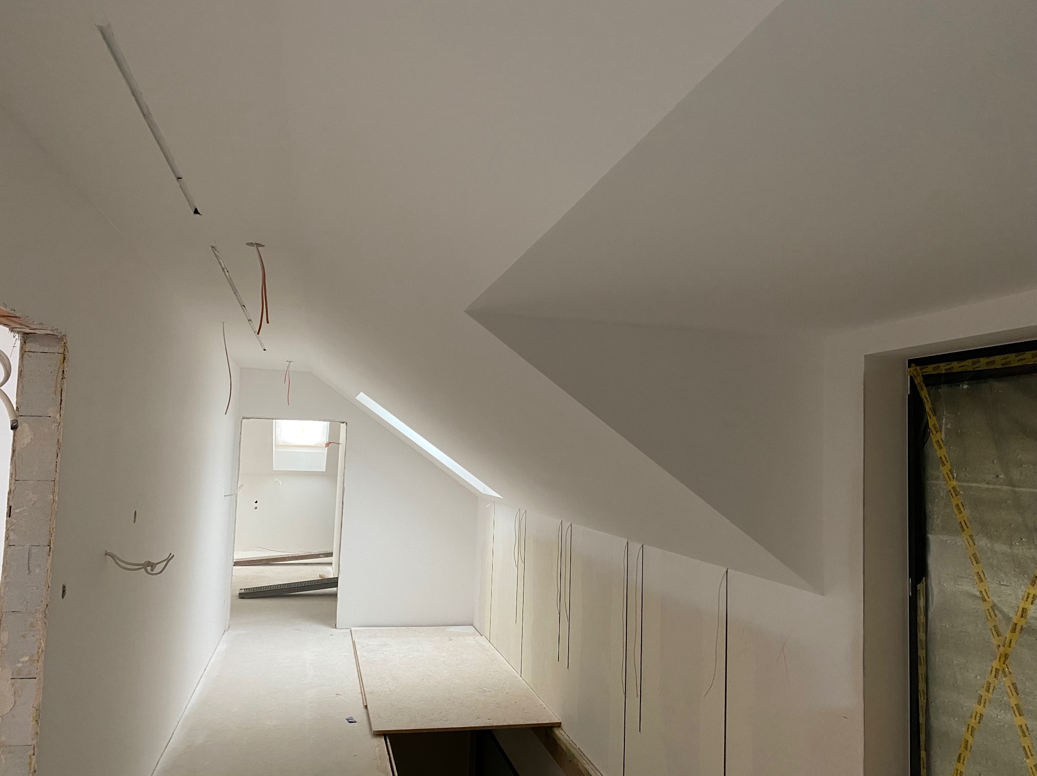 Poddasze w trakcie remontu: białe ściany, skośny sufit, widoczne przewody elektryczne, fragment otworu w podłodze, zabezpieczone folią drzwi.
