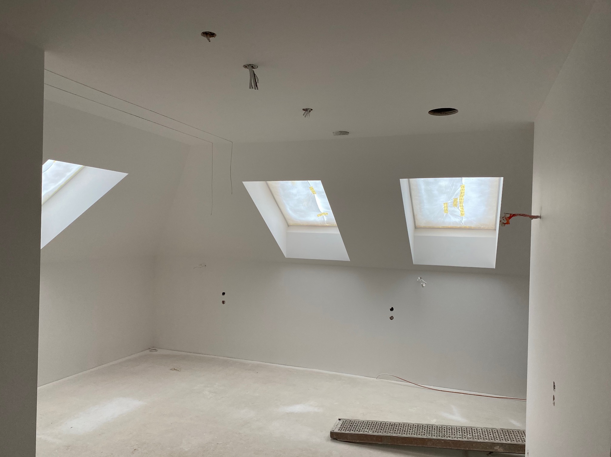 Poddasze w trakcie prac wykończeniowych, widoczne trzy okna dachowe, gładkie, białe ściany i sufit z wystającymi przewodami elektrycznymi, na podłodze leży metalowa kratka.