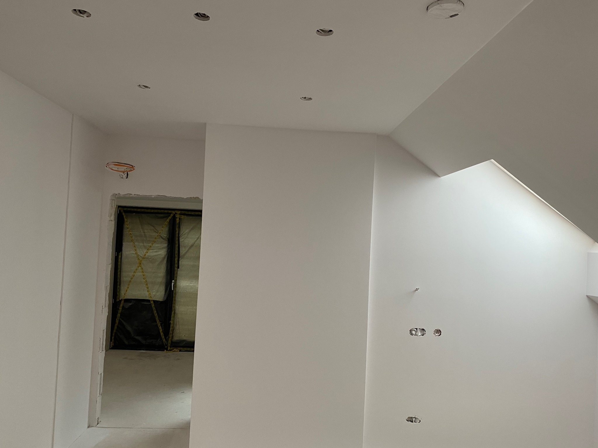 Wnętrze w trakcie malowania na biało, widoczne otwory na oświetlenie w suficie, niezamaskowane gniazdka elektryczne w ścianie, drzwi zabezpieczone folią ochronną.