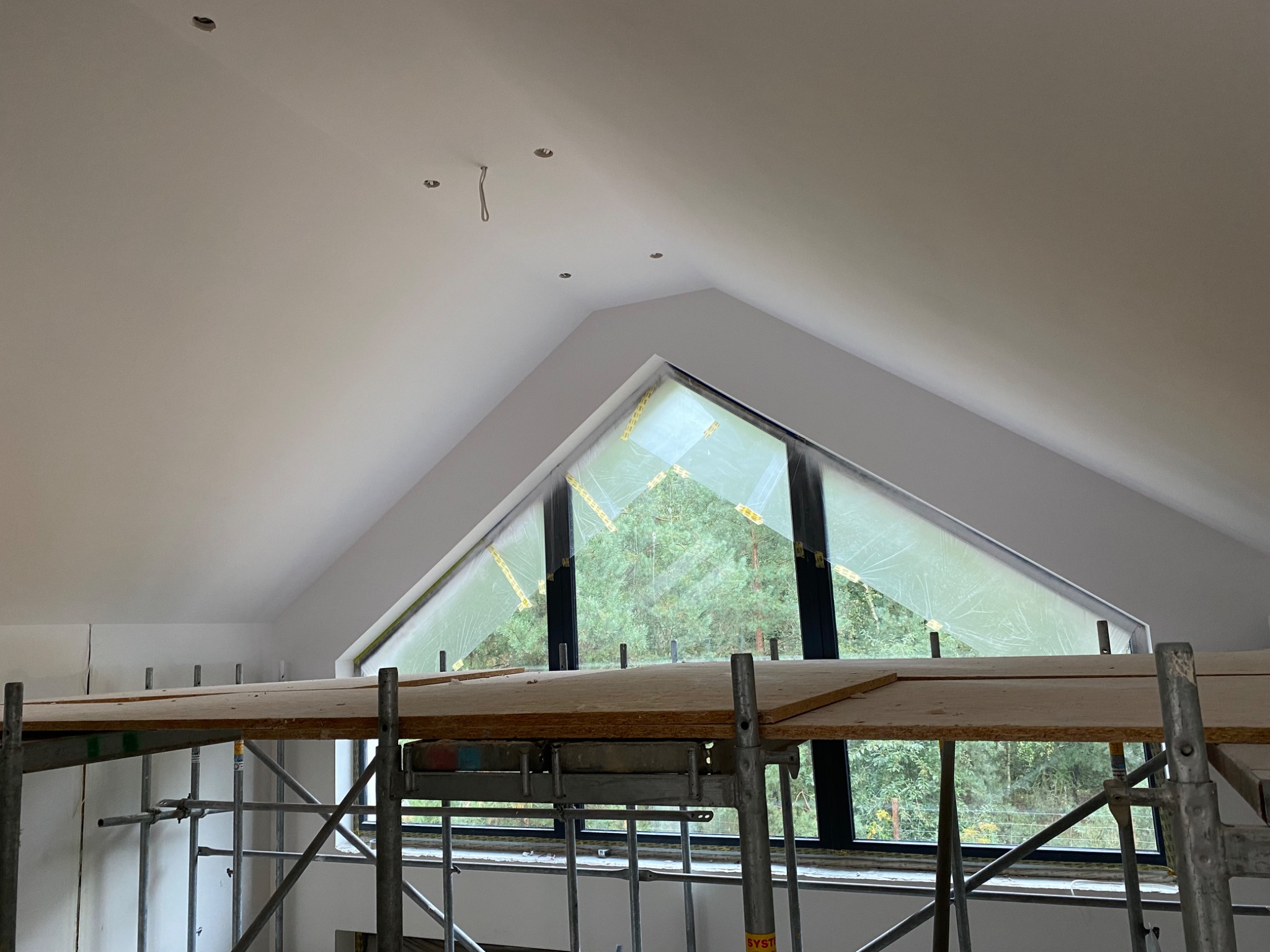 Poddasze z oknem w kształcie trójkąta zabezpieczonym folią, widoczne rusztowanie i otwory na oświetlenie w świeżo pomalowanym suficie.