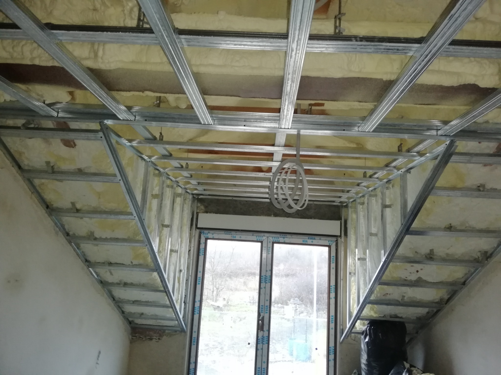 Szkielet konstrukcji sufitu podwieszanego z metalowych profili, wypełniony pianką izolacyjną, widok na okno w skosie poddasza.
