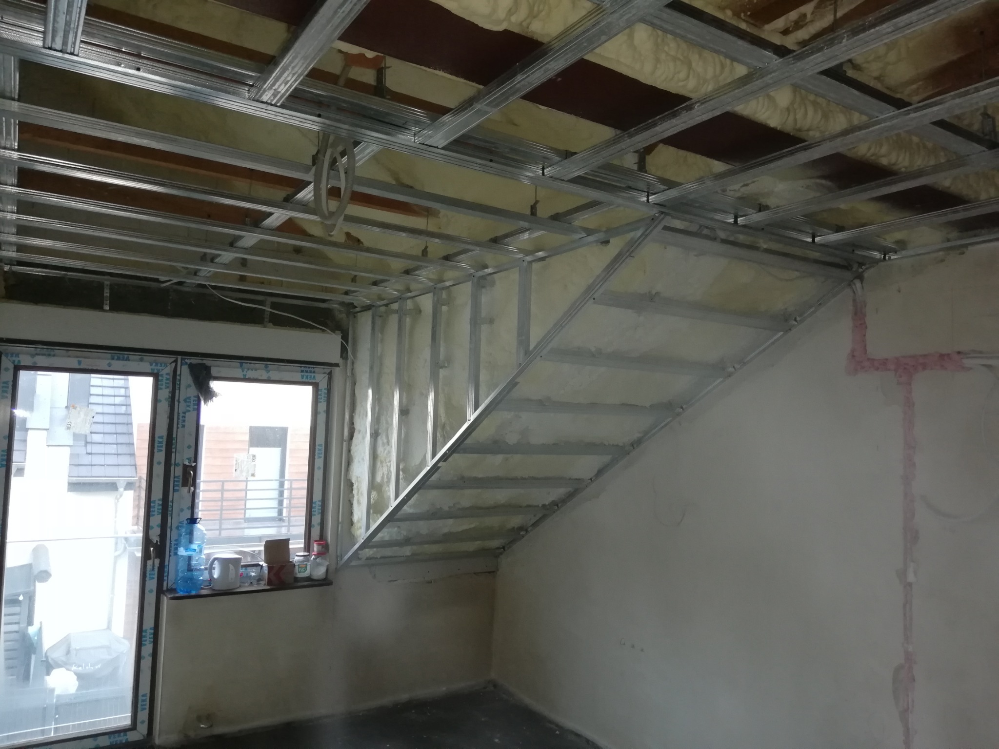 Wnętrze w trakcie remontu z widoczną konstrukcją metalową sufitu podwieszanego i ścianki działowej, ociepleniem z wełny mineralnej oraz fragmentem okna z butelkami i kubkami na parapecie.