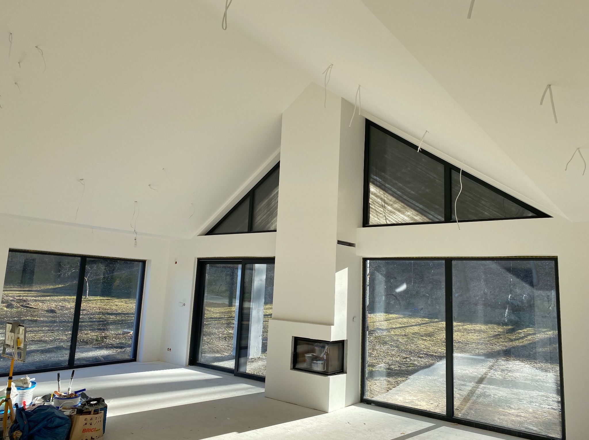 Jasne wnętrze z dużymi oknami i kominkiem, świeżo po malowaniu. Widoczne narzędzia malarskie sugerują etap wykończeniowy. Minimalistyczny design z elementami surowego betonu.