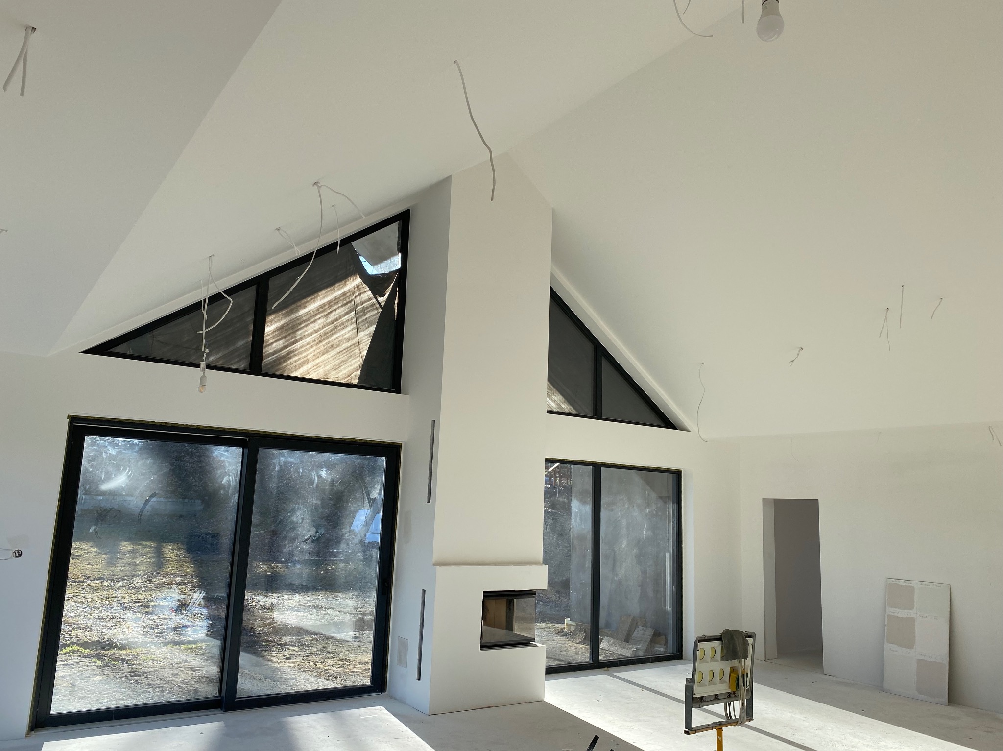 Jasne wnętrze z dużymi oknami i kominkiem, w trakcie wykańczania. Widoczne wiszące kable, próbki kolorów na ścianie i narzędzia malarskie. Minimalistyczny design.